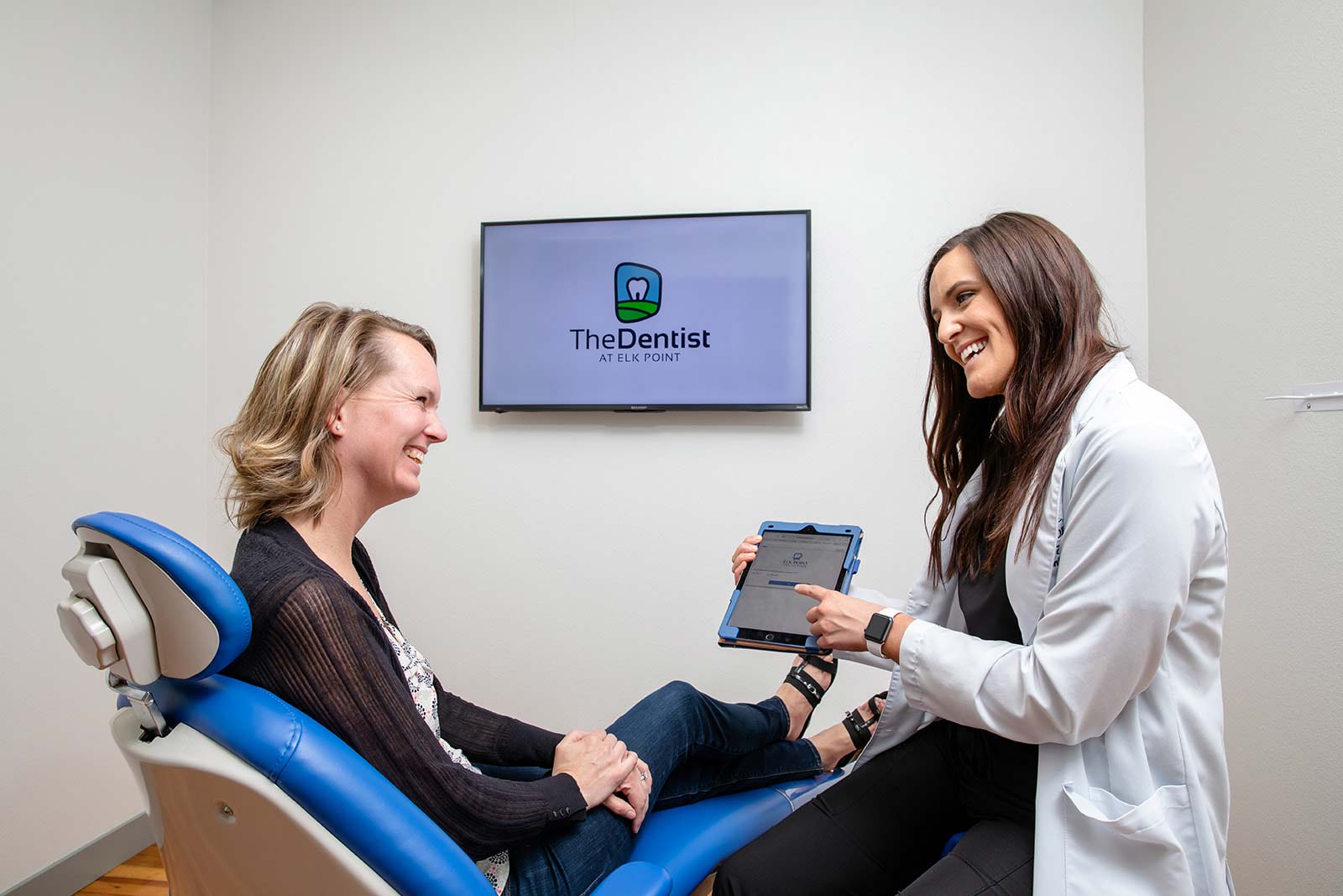 A dentist holding an iPad is talking to a dental patient who is seated in an exam chair
